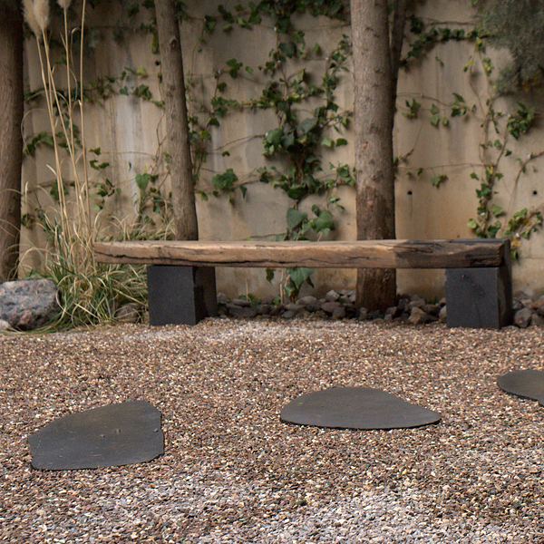 A reclaimed timber beam bench on two black basalt blocks, against a vine-covered wall, with stepping stones in the gravel in front.