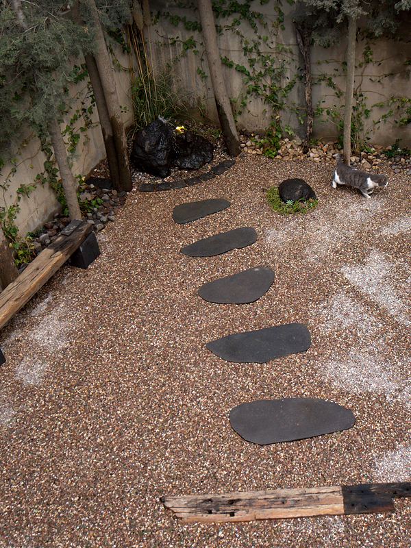 The gravel courtyard seen from above: a curve of basalt stepping stones, a timber beam bench, a mossy black rock and a cat crossing the gravel.