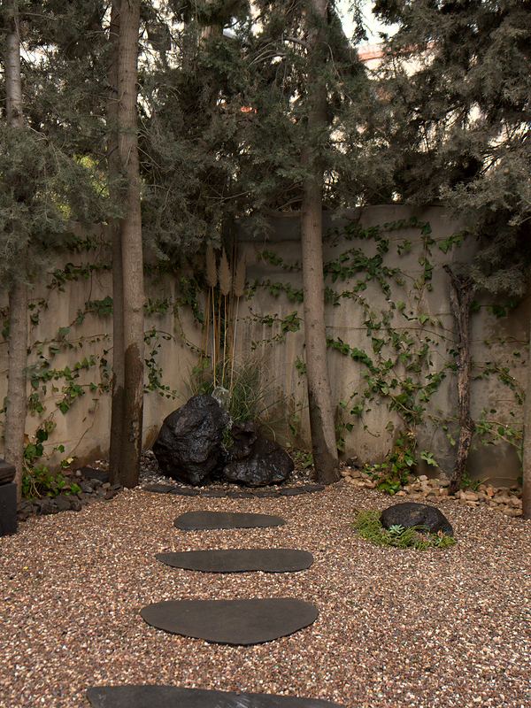A cluster of black volcanic boulders and pampas grass under the cypresses, with basalt stepping stones leading to them through gravel.