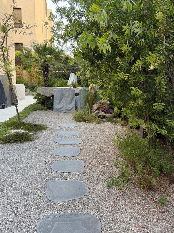 Dark stepping stones leading through gravel towards a stone table under palms.